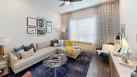 This single-floor residence showcases a thoughtfully designed open-concept layout with contemporary finishes throughout. The kitchen serves as a standout feature, offering an expansive white-quartz island with bar seating, dark espresso shaker cabinetry extending to the ceiling, and a full suite of stainless steel appliances including a French door refrigerator, electric range, and built-in microwave. Under-cabinet lighting illuminates the stone tile backsplash, while wood-look flooring ties the space together seamlessly.

The living area is bright and welcoming, benefiting from large windows dressed with patterned drapes that invite generous natural light. A ceiling fan adds functional comfort to the relaxed atmosphere.

The bedrooms feature plush carpeting, ceiling fans, and sizable windows with outdoor views, creating a calm and restful environment. Ample doorways suggest well-considered spatial flow between rooms.

The bathroom is finished with horizontally laid tile surrounding a soaking tub and shower combination, accented by a decorative mosaic border. A dark-toned vanity with white quartz countertop and wood-look tile flooring complete the refined, cohesive aesthetic found throughout this well-appointed residence.