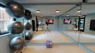 This single-level fitness studio is thoughtfully designed to support a wide range of exercise activities, from yoga and Pilates to group fitness classes and personal training sessions. The room is finished in a calming light blue-gray palette, creating an open and motivating environment. Light wood-toned flooring runs throughout, providing a clean, low-impact surface well-suited for floor exercises and movement-based workouts.

Floor-to-ceiling mirrors span a full wall, maximizing the sense of space while allowing users to monitor their form during exercise. Two wall-mounted flat-screen televisions are positioned for easy viewing from anywhere in the room, supporting instructor-led or virtual workout sessions. A smart thermostat and integrated HVAC vents ensure comfortable climate control year-round.

Equipment storage is conveniently integrated into the studio, with a multi-tiered rack holding a full set of stability and exercise balls in varying sizes. Wall-mounted exercise mats are accessible for immediate use. Natural light filters into the space through a window offering a view of the surrounding streetscape, adding a welcoming connection to the outdoors.

A glass-paneled door connects directly to an adjacent fitness center equipped with cardio and strength training machines, making this studio a seamless part of a larger, comprehensive fitness facility.