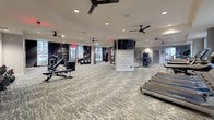 This expansive fitness center is thoughtfully designed to accommodate a full range of workout routines across multiple clearly defined zones. The main floor is covered in a durable, textured carpet and illuminated by recessed lighting and multiple ceiling fans that maintain a comfortable environment throughout. Large windows line the perimeter, inviting generous natural light and providing open views of the surrounding area.

The cardio zone features a row of modern treadmills equipped with individual screens, positioned directly in front of floor-to-ceiling windows. Nearby, upright bikes and elliptical machines offer additional cardio options. The strength training area is comprehensively outfitted with a multi-functional cable and suspension training rig, a full rack of dumbbells displayed in front of large wall mirrors, selectorized weight machines, adjustable benches, and stability balls.

Motivational murals and bold typographic wall graphics add energy and personality to the space. A separate studio room with hardwood flooring and mirrored walls provides an ideal setting for group classes, stretching, or mat-based workouts. The fitness center also includes restroom facilities and ample open floor space, ensuring a comfortable and functional experience for users of all fitness levels.