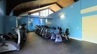 This fitness center offers an inviting and functional space designed to support a well-rounded workout routine. The room is defined by its striking architectural character, most notably the dramatic vaulted ceiling with exposed wood paneling that rises to a generous peak, lending the space an open, airy quality rarely found in typical gym settings. Clerestory windows positioned near the roofline allow natural light to filter in, brightening the interior and creating a connection to the surrounding greenery visible outside.

The walls are painted a bold, energizing shade of blue, contributing to a motivating atmosphere throughout. Rubber flooring runs across the entire space, providing a durable and practical surface suited for high-traffic fitness use. The cardio equipment area is well-stocked, featuring a row of treadmills, recumbent bikes, and additional cardio machines, all positioned to maximize the use of available floor space. Toward the back wall, a weight rack and strength training equipment are visible, rounding out the facility's offerings.

Pendant lighting and ceiling fans complement the natural light, ensuring the space remains comfortable during all hours of use. Overall, the fitness center combines functional equipment with distinctive architectural charm.