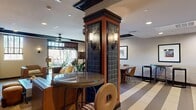 Located at 4108 Del Rey Ave in Marina del Rey, Los Angeles, this thoughtfully designed single-floor space offers a seamlessly open layout that blends multiple functional zones into one cohesive environment. The interior is anchored by a fully equipped kitchen featuring dark espresso cabinetry, stainless steel appliances including a full-size refrigerator and dishwasher, granite countertops, a mosaic tile backsplash, and recessed lighting overhead. A striped large-format tile floor runs throughout, adding a bold graphic element that visually defines the space. A distinctive tiled column with warm wall sconces serves as an architectural focal point, separating the kitchen and dining areas from the lounge zone. The lounge area is furnished with rich cognac leather armchairs and anchored by large windows dressed with wood blinds, filling the room with generous natural light and views of lush outdoor greenery. Dark wood crown molding and ceiling beams add depth and sophistication. Pendant lighting clusters and accent lamps contribute to a warm, layered ambiance throughout. The dining area accommodates small group seating, while framed artwork and live potted plants enhance the overall aesthetic. The space strikes a balance between modern amenities and warm, inviting design details.