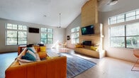 This single-floor community space is designed for socializing and recreation, combining open-concept living with functional amenities. The centerpiece of the main gathering area is a striking floor-to-ceiling light brick fireplace, topped with a wall-mounted television and flanked by a built-in bench with cushioned seating. Soaring vaulted ceilings amplify the sense of openness, while large multi-pane windows on multiple walls invite abundant natural light and views of the surrounding greenery. Light wood-tone flooring runs throughout, lending warmth and visual continuity across the space. The lounge area accommodates ample seating, and a high-top table with wire-frame chairs provides a casual dining or conversation nook near the windows. A glass door leads to an outdoor deck, extending the living space further. The kitchen is well-appointed with stainless steel appliances — including a full-size refrigerator, electric range with a wall-mounted range hood, and microwave — set against clean white cabinetry with wood-toned upper cabinets. A breakfast bar with vibrant orange wire barstools adds a playful, modern touch. Adjacent to the main lounge, a spacious game room features vaulted ceilings, a foosball table, and a wall-mounted TV, making it an ideal space for entertainment and leisure.