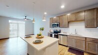 Located at 1850 Summerfield Way in Sun Prairie, Wisconsin, this single-floor residence offers a thoughtfully designed open-concept layout with modern finishes and a clean, contemporary aesthetic. The kitchen serves as the heart of the home, showcasing warm-toned shaker-style cabinetry, a white quartz waterfall island with pendant lighting overhead, a classic subway tile backsplash, and a full suite of stainless steel appliances including a range, microwave, dishwasher, and refrigerator. Light hardwood-style flooring flows seamlessly throughout the main living areas, enhancing the bright and airy feel of the space. The living area features a ceiling fan and a large sliding glass door framed by vertical blinds, allowing abundant natural light to fill the room. The bedroom offers plush carpeting, a ceiling fan, and a window with neighborhood views, creating a comfortable and restful retreat. The bathroom is finished with white cabinetry, a wide vanity with an undermount sink, a wood-framed mirror, and warm lighting. Clean neutral walls and consistent flooring choices throughout the home contribute to a cohesive, move-in-ready atmosphere suited for modern living.