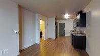This single-floor unit offers an open-concept layout that efficiently combines the living and kitchen areas into one cohesive space. The warm hardwood-style flooring runs throughout, lending a consistent and inviting character to the interior. Crisp white walls and smooth ceilings keep the atmosphere bright and clean, complementing the overall modern, functional design.

The kitchen is positioned along one wall and features dark cabinetry, granite-style countertops, stainless steel appliances including a full-size refrigerator and microwave, and a sink — providing all the essentials within a well-organized footprint. The open living area adjacent to the kitchen offers flexible space for furnishing and everyday use.

A full bathroom is accessible directly from the main living area and includes a toilet, vanity with sink, and a shower, finished in warm-toned lighting that creates a comfortable atmosphere. The front entry door is centrally situated, offering straightforward access to the main living space.

Overall, the unit is designed with practicality and efficiency in mind, making thoughtful use of its single-floor layout. The cohesive flooring, neutral color palette, and modern fixtures combine to create a clean, move-in-ready environment suited for everyday living.