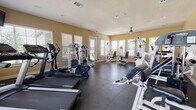 This fitness center offers a comprehensive and well-organized workout facility designed to accommodate a wide range of exercise routines. The space is laid out on a single floor, making it easily accessible and straightforward to navigate.

The room is bathed in natural light, courtesy of numerous large windows fitted with white horizontal blinds positioned along multiple walls. The warm, neutral-toned walls and recessed lighting create a bright and welcoming atmosphere throughout.

The flooring consists of dark, durable rubber matting — a practical and safety-conscious choice for a high-use exercise environment. Cardio equipment is well-represented, with multiple treadmills, including Landice and Galaxy Promaxima models, alongside elliptical trainers and recumbent stationary bikes by SportsArt.

The strength training section is equally thorough, featuring several cable and weight machine stations suitable for a variety of muscle groups, including chest press, leg press, and lat pulldown configurations. A flat utility bench accompanies the free weight area for added versatility.

Overhead, ceiling fans assist with air circulation, and a wall-mounted television provides entertainment during workouts. An informational bulletin board is also visible near one of the entrance doors. Multiple entry and exit points, including a clearly marked emergency exit, ensure safe and convenient access throughout the facility.