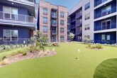 Located in Allen, Texas, this thoughtfully designed outdoor amenity space serves as a central gathering hub for its surrounding multi-story residential community. The complex showcases a striking contemporary architectural style, combining warm brick facades with bold blue and neutral panel cladding, multiple private balconies, and large windows that overlook the shared grounds below.

At the heart of the space is a generously sized putting green with lush artificial turf, multiple pin placements, and organically shaped landscaped islands featuring ornamental grasses, native shrubs, flowering plants, and natural boulders. The undulating layout of the green provides an engaging recreational experience suitable for all skill levels.

Adjacent to the putting green is a full pickleball and tennis court finished in a vibrant teal and green surface, enclosed by metal fencing with gated entry and outfitted with courtside seating benches. Overhead string lighting and pole-mounted fixtures add evening ambiance throughout the common walkways.

Connecting these amenities is a clean concrete pathway lined with drought-tolerant Texas-friendly landscaping, including ornamental yuccas and seasonal color plantings. The overall atmosphere is active yet inviting, offering residents well-maintained outdoor recreation facilities under open Texas skies.