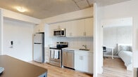 Located at 575 Princess St in Kingston, Ontario, this modern 3-bedroom, 2-bathroom unit showcases a contemporary industrial aesthetic throughout. Exposed concrete ceilings contrast beautifully with crisp white walls and light hardwood flooring, creating a clean, urban ambiance across the single-floor layout.

The open-concept kitchen and living area form the heart of the unit. The kitchen is well-appointed with stainless steel appliances — including a refrigerator, electric range, built-in microwave, and dishwasher — complemented by white cabinetry, a subway tile backsplash, and a double sink. A bar-height dining table connects the kitchen to the living area, which features a large window that brings in natural light and outdoor views. In-unit washer and dryer are conveniently tucked into a dedicated laundry alcove nearby.

The primary bedroom is a comfortable, well-proportioned retreat with a large window overlooking the surroundings, roller blinds for privacy, and light hardwood floors. The additional bedrooms also feature hardwood flooring and exposed concrete ceilings, maintaining the unit's cohesive modern style. The two bathrooms are finished with white subway tile surrounds, soaker tubs, and clean, minimalist fixtures, offering a functional and polished finish throughout.