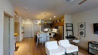 This single-floor residence in Euless, TX, showcases a thoughtfully designed open-concept layout that seamlessly connects the kitchen, dining, and living areas. The kitchen is a standout feature, equipped with stainless steel appliances — including a full-size refrigerator, microwave, and range — complemented by rich dark wood cabinetry that extends to the upper walls, where decorative plants add a natural accent. Light-colored countertops provide a clean contrast, and a peninsula with bar-height seating offers a casual dining option beneath a trio of modern pendant lights. Recessed lighting throughout keeps the space bright and well-illuminated. Warm-toned hardwood flooring runs continuously across the open floor plan, enhancing the cohesive and inviting atmosphere. The living area accommodates plush white seating and is accented by a sleek ladder-style shelving unit. A clean entryway with a solid door is visible from the main living space, contributing to the unit's practical, streamlined design. The overall aesthetic balances modern minimalism with warm, residential comfort.