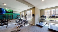 This fitness center offers a comprehensive, single-floor workout facility designed to support a wide range of exercise routines. The space is visually defined by its bold teal accent walls paired with neutral taupe tones, creating an energetic yet focused atmosphere. Durable patterned carpet tiles cover the floor throughout, providing a practical surface suited for both cardio and strength training activities.

The cardio zone features multiple treadmills, elliptical machines, and stationary bikes, all positioned facing large, multi-pane windows that bring in ample natural light and offer views of the surrounding exterior. Full-length mirrors line one wall, enhancing the sense of space and allowing users to monitor their form during workouts.

The strength training area is well-appointed with a full dumbbell rack offering a range of weights, adjustable workout benches, and a multi-functional cable machine system suitable for a variety of resistance exercises. Additional strength equipment, including a back extension bench and other targeted machines, occupies a dedicated section of the facility.

A large flat-screen television mounted on the mirrored wall adds an entertainment element to the space. Recessed and flush-mount ceiling lighting ensures the facility is well-illuminated throughout. The fitness center is accessible via a glass-paneled entry door, and climate control is managed through visible wall-mounted thermostats and ceiling ventilation.