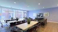 This single-floor community space is thoughtfully designed to serve as both a social hub and a comfortable retreat. The room is finished with soft lavender walls and a clean white ceiling punctuated by recessed lighting, creating a bright and inviting ambiance throughout. Large floor-to-ceiling windows along one wall flood the interior with natural light, offering views of the surrounding brick exterior and lush greenery outside.

The layout is divided into distinct functional zones. A dining area features multiple rectangular white tables paired with dark modern chairs, each table accented with potted greenery that adds a touch of nature to the setting. Complementing this is a lounge area furnished with upholstered armchairs and a sofa arranged around a wall-mounted flat-screen television, providing a comfortable space for relaxation or casual conversation. A media console and sideboard nearby offer practical storage and display surfaces.

The flooring transitions subtly across the space, and the overall design strikes a balance between practicality and comfort. Decorative elements, including framed artwork and carefully placed plants, contribute to the welcoming atmosphere. This versatile, well-appointed space is well-suited for dining, socializing, and everyday community life.