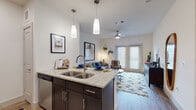 This single-story unit offers a thoughtfully designed open-concept layout that seamlessly connects the kitchen, dining, and living areas. The kitchen is outfitted with dark espresso cabinetry, granite countertops, a tiled backsplash, and a full suite of stainless steel appliances, including a range, over-the-range microwave, dishwasher, and refrigerator. A central island with a double basin sink and pendant lighting above serves as both a functional workspace and a natural dividing point between the kitchen and living area. Wood-look flooring runs throughout the main living spaces, adding warmth and continuity. The living room is bright and welcoming, featuring a ceiling fan, large windows with blinds, and a glass door that allows natural light to filter in. The bedroom is carpeted and comfortably sized, offering a walk-in closet and a window for natural light. The full bathroom includes a tiled tub-shower combination with a curved curtain rod, wood-look flooring, and clean, neutral finishes. The entryway provides convenient access alongside ample closet storage. Throughout the unit, crisp white walls and modern fixtures create a clean, contemporary aesthetic well-suited for comfortable everyday living.