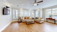 Located at 201 Kramer Ct in Greer, South Carolina, this single-floor space offers a welcoming and well-appointed living area designed for both comfort and functionality. The open-plan layout seamlessly connects the living room to an adjacent kitchen area, which features white cabinetry, a breakfast bar with bar seating, and black appliances — all visible through a wide pass-through opening framed by a decorative column.

The living room itself is generously proportioned, finished with warm hardwood-style flooring that runs throughout and is anchored by a neutral color palette of soft grays and crisp white trim. Crown molding adds an elegant architectural detail along the ceiling line, while recessed lighting provides even, ambient illumination. A ceiling fan with an integrated light fixture ensures year-round comfort.

Natural light is a defining feature of this space, with an expansive bank of windows along two walls — complete with transom windows above — flooding the interior with daylight and offering views of the surrounding neighborhood. The bright, airy ambiance is further enhanced by the light-toned walls and open sightlines throughout. The space also includes a convenient exterior access door. Overall, the design strikes a balance between casual livability and thoughtful detailing, making it a comfortable and inviting environment.