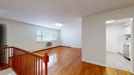 This single-floor space showcases a clean, functional design highlighted by rich hardwood flooring that flows continuously through the main living areas and bedrooms, lending warmth and cohesion to the interior. The open living area is generously proportioned, featuring a staircase landing with a classic wood railing, a flush-mount ceiling light, and a window fitted with blinds that admits natural light. Baseboard heating runs along the perimeter walls throughout, ensuring efficient climate control, while wall-mounted air conditioning units in select rooms provide additional comfort.

The kitchen is efficiently designed with white shaker cabinetry, a white subway tile backsplash, light luxury vinyl plank flooring, a stainless steel sink with a matte black pull-down faucet, and stainless steel appliances including a microwave and range. A pass-through opening connects the kitchen to the main living space, enhancing the sense of openness.

The bedrooms are bright and well-sized, each benefiting from windows with blinds and baseboard heating. Closet space is well-distributed, with walk-in and standard closets featuring built-in shelving and matte black hardware. The bathroom is finished with large-format marble-look wall tiles, a white soaking tub, and matte black fixtures, creating a refined, contemporary aesthetic throughout.