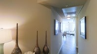 This single-story space is defined by a well-appointed hallway that serves as the central passageway connecting the entry to the main living area. The corridor is finished with warm beige walls and smooth hardwood-style flooring that extends throughout, creating a cohesive and inviting aesthetic. Decorative tall vase accents in a hammered bronze finish add an artistic touch near the entry, complementing the neutral color palette. Framed wall art lines the hallway, contributing to the curated, polished atmosphere of the space.

The hallway opens into a bright and airy living area, where a ceiling fan with a light fixture provides both airflow and ambient illumination. The living space is furnished with comfortable seating arrangements and soft area rugs, suggesting a well-organized and functional layout. Recessed lighting and flush-mount ceiling fixtures ensure even illumination throughout. The transition from the intimate corridor to the more expansive living area creates a pleasant sense of arrival. Overall, the space combines modern design sensibilities with warm, neutral finishes to create a comfortable and visually cohesive environment.