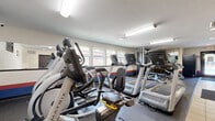 Americana's Gym is a single-floor exercise facility located at 12598 W Dakota Ave in Lakewood, Colorado. The space is efficiently organized to maximize its compact footprint, offering a focused and functional workout environment. The room features durable rubber flooring throughout the main exercise area, transitioning to light tile near the entryway, and is illuminated by fluorescent strip lighting complemented by recessed and track fixtures that keep the space bright and practical. A row of Matrix-brand treadmills lines one wall, accompanied by an elliptical trainer and a stationary recumbent bike, providing a comprehensive cardio selection. A multi-functional cable strength machine anchors the center of the room, while a set of medicine balls on a wall-mounted rack offers additional training variety. A padded workout bench adds further functionality for strength exercises. Windows along the side wall allow natural light to filter in and offer views of the surrounding Colorado landscape. The walls are decorated with a bold red, white, and blue horizontal stripe accent, reinforcing the gym's patriotic branding. A motivational typographic wall mural and an "Americana's Gym" sign add character and energy to the space. A wall-mounted television provides entertainment during workouts. The room is accessed through a dedicated entry door with a key access panel, ensuring a secure and private fitness environment.