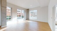 Located at 2065 South River Parkway in Portland, Oregon, this single-floor unit offers a clean, contemporary design throughout. Light-toned hardwood flooring flows seamlessly through the open-concept living and kitchen areas, creating a cohesive and airy feel. The living area is a standout space, featuring a striking corner arrangement of large windows that flood the room with natural light and frame views of the surrounding Portland cityscape, including neighboring brick buildings and lush greenery. An exposed concrete pillar adds an industrial architectural accent to this bright, open room.

The kitchen is thoughtfully laid out with warm wood cabinetry extending to the ceiling for ample storage, a white tile countertop peninsula with an integrated sink, and white appliances including a refrigerator, electric range, and dishwasher. Recessed lighting above enhances the clean, functional aesthetic.

The bedroom is a cozy, carpeted retreat with a tall window that invites natural light and offers views of the urban surroundings. The bathroom features a full soaking tub and shower combination with chrome fixtures, set against crisp white walls and neutral flooring. Throughout, the space maintains a bright, minimalist character suited to urban living.