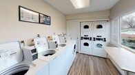 The Olympus Park Laundry Facility is a thoughtfully designed shared laundry room that offers residents a clean, functional, and comfortable space for their laundry needs. The room is outfitted with a row of commercial-grade top-load and front-load washers, including Speed Queen models, arranged along one wall for easy access. Stacked dryer units line the adjacent wall, maximizing the use of vertical space and providing ample drying capacity. The flooring throughout is a durable wood-look laminate that is both practical and visually appealing. A large window along one wall fills the room with natural light and offers views of the surrounding outdoor landscaping and parking area, creating a bright and pleasant atmosphere. A fold-down countertop shelf beneath the window provides a convenient surface for folding and sorting laundry. The room also features a vending machine for laundry supplies, an electrical panel box, and posted informational signage on the walls. Access to the facility is through a glass-paneled door clearly marked with the Olympus Park Laundry Facility logo. The overall layout is clean, organized, and efficiently designed to serve the needs of residents with ease.
