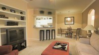 This single-floor space is designed with an open-concept layout that promotes a natural flow between the living, kitchen, and dining areas. The living room features plush carpeting, a built-in shelving unit ideal for displaying décor, and a cozy corner accommodating an entertainment setup. Crown molding runs throughout, adding a refined architectural touch to the warm, neutral-toned walls.

The kitchen is accessed via a stylish pass-through bar, complete with dark bar stools that provide casual seating. White cabinetry lines the kitchen walls, complemented by open upper shelves adorned with potted greenery, lending a fresh, lively accent to the space. The kitchen is well-lit and efficiently laid out, with appliances visible through the pass-through opening.

Adjacent to the kitchen, the dining area is defined by a chair-rail molding detail and anchored by a round glass-top table surrounded by elegant wrought-iron chairs. A pendant light fixture hangs above, creating an intimate atmosphere, while a framed piece of artwork on the wall adds a decorative focal point. Together, the interconnected spaces create a cohesive and inviting environment well-suited for both everyday living and entertaining.