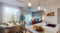 This thoughtfully designed single-floor residence combines modern aesthetics with comfortable living across a well-connected open-concept floor plan. The kitchen is outfitted with light wood cabinetry, granite countertops, stainless steel appliances, and track lighting, flowing seamlessly into a dining area distinguished by a bold slate-blue accent wall, a round dining table with velvet chairs, and a large decorative woven wall piece. Natural light pours in through oversized windows dressed with patterned drapes. The living room continues the contemporary theme with hardwood-style flooring, a striking large-scale abstract artwork above the sofa, and a sliding glass door that opens to an outdoor area, filling the space with additional light and views of greenery. Two bedrooms offer plush carpeting, ceiling fans, and generous windows. The primary bedroom features a dramatic teal velvet headboard against a deep accent wall, while the secondary bedroom offers a warmer, neutral palette with a mid-century dresser. The bathroom presents a clean, modern design with light wood vanity cabinetry, granite countertops, a large mirror, and wood-look tile flooring. Throughout the residence, cohesive design elements and quality finishes create an inviting, contemporary ambiance.
