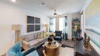 This single-floor residence offers a cohesive and contemporary living experience with a warm, curated aesthetic throughout. The living room serves as the heart of the home, featuring hardwood-style flooring, a ceiling fan, and a large window dressed in teal tie-dye curtains that invite natural light. A neutral sofa accented with colorful throw pillows, a sculptural gold floor lamp, and a set of four textured black wall art panels give the space a modern yet inviting character. An open shelving unit adds functional display space near the window. The kitchen is well-equipped with dark shaker-style cabinetry, stainless steel appliances including a refrigerator, range, and microwave, a subway tile backsplash, and a generous island with a white quartz countertop and undermount sink — ideal for both cooking and casual dining. The bedroom is carpeted and comfortably sized, featuring a ceiling fan, a large window with gold drapery, and ample wall space for artwork. It connects directly to a bathroom that boasts dark cabinetry, a long white vanity countertop with dual sinks, and a walk-in closet with built-in shelving. The overall palette of teal, gold, and warm neutrals creates a polished, cohesive ambiance throughout the residence.