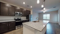Located at 3680 Avalon Park E Blvd in Orlando, Florida, this single-floor unit offers a thoughtfully designed open-concept layout that seamlessly connects the kitchen, dining, and living areas. The kitchen serves as the centerpiece of the space, showcasing rich dark espresso cabinetry paired with speckled granite countertops that extend across a generously sized center island equipped with a built-in sink and pendant lighting overhead. A full suite of stainless steel appliances—including a refrigerator, electric range, over-the-range microwave, and dishwasher—completes the kitchen's functional and modern aesthetic. Light wood-style flooring runs throughout the main living areas, providing a warm and cohesive look, while recessed lighting and ceiling fans ensure consistent illumination and comfort. The open living area benefits from natural light admitted through windows fitted with horizontal blinds. The bedroom features darker hardwood-style flooring, a ceiling fan, and a window that brings in additional daylight. The bathroom is finished with matching dark cabinetry, granite countertops, wood-style flooring, and a tiled tub-shower combination, maintaining the consistent design language found throughout the unit.
