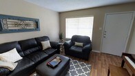 This single-floor residence offers a comfortable and functional layout designed for everyday living. The living room serves as the heart of the space, featuring warm wood-look hardwood flooring that adds a sense of character and continuity throughout the area. A large window fitted with horizontal blinds allows abundant natural light to fill the room, creating a bright and inviting atmosphere. The textured ceiling adds a classic touch to the overall aesthetic. The living space is well-proportioned, comfortably accommodating a full seating arrangement that includes a sofa, an oversized armchair, and an ottoman, all centered around a patterned area rug that anchors the room. Decorative wall accents and potted greenery contribute to the homey ambiance, while neutral wall tones provide a versatile backdrop that complements a variety of furnishing styles. The entry door is conveniently accessible from the living area, and the overall layout reflects a practical, easy-to-navigate floor plan. This space is well-suited for those seeking a straightforward, single-level living environment with a warm and welcoming feel.