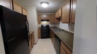 This single-floor residence offers two bedrooms and one and a half bathrooms, designed with practicality in mind. The kitchen follows a classic galley layout, maximizing efficiency within a defined corridor-style space. Warm-toned wooden cabinetry lines both walls from floor to ceiling, providing generous storage throughout. Dark countertops run the length of the kitchen on both sides, offering ample prep space. Black appliances, including a refrigerator, electric range, and dishwasher, create a cohesive, uniform look. An under-cabinet range hood is positioned above the stove, while a double-basin sink with a standard faucet is conveniently situated along the opposite counter. The light-toned flooring contrasts with the darker countertops and appliances, keeping the space feeling bright. A flush-mount ceiling light provides overhead illumination. The textured ceiling adds a subtle architectural detail consistent throughout. The overall layout is straightforward and functional, making efficient use of available space while providing all essential kitchen amenities needed for comfortable everyday living.