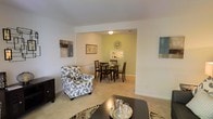 This two-story home offers a welcoming interior defined by clean lines, neutral color palettes, and a seamlessly connected layout. The main living area features plush wall-to-wall carpeting and crisp white walls adorned with framed artwork, creating a calm and inviting atmosphere. A comfortable seating area anchors the living space, centered around a sleek dark coffee table that adds a modern contrast to the soft surroundings.

The living area flows naturally into a dedicated dining space, distinguished by a warm sage-green accent wall that adds a subtle pop of color. A circular dining table surrounded by dark wood chairs occupies this area, making it well-suited for everyday meals or entertaining guests. A decorative starburst wall clock serves as a focal point above a small wall-mounted shelf, adding character to the space. Ceiling-mounted lighting provides consistent illumination throughout both areas.

The open-plan arrangement between the living and dining zones enhances the sense of spaciousness, encouraging easy movement and social interaction. The second floor further expands the living options within this home, offering additional versatility and space across two well-considered levels.