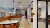 Located at 3421 N Freeway Blvd in Sacramento, CA, this thoughtfully designed single-floor commercial space offers a sophisticated blend of modern aesthetics and warm, natural materials. Light wood-look tile flooring flows seamlessly throughout, unifying the various functional areas. The two living rooms provide versatile lounge settings, one featuring rich walnut built-in shelving, eclectic accent seating, and a decorative metallic chain-link room divider that adds visual depth. A curated workstation area displays striking large-format abstract artwork against a textured brick-style wall, flanked by sliding barn doors in a deep navy finish. The dining room is a standout feature, lined with large windows that invite natural light and anchored by a long wood conference-style table surrounded by plush velvet seating beneath cascading gold pendant lights. The private office, accessible via a sleek black-framed sliding glass door, is efficiently outfitted with a built-in desk and ample storage. Vertical wood slat partitions and booth-style seating with marble-top tables add warmth and structure throughout the hallway and transitional spaces. The overall ambiance is polished yet inviting, with a cohesive palette of walnut wood tones, warm metallics, and crisp white walls.