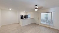Located at 1405 W North Loop Blvd in Austin's 78756 zip code, this single-floor unit offers a clean, modern aesthetic throughout its open-concept layout. Light wood-tone vinyl flooring runs continuously across the living and kitchen areas, creating a cohesive and airy feel. The kitchen is outfitted with white cabinetry extending to the ceiling for ample storage, a subway tile backsplash, and black appliances including a refrigerator, microwave, and range. A peninsula counter with a white surface separates the kitchen from the living space, providing a functional bar-style transition between the two areas. The living room benefits from a ceiling fan with integrated lighting and a large window that invites natural light while offering views of the outdoors. White walls and a textured ceiling reinforce the bright, neutral palette throughout. A hallway leads to additional rooms, maintaining a straightforward and functional floor plan. The unit's fresh, updated finishes and single-level layout make it a practical and well-appointed space in central Austin.