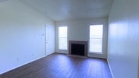 This single-floor space offers a clean, functional layout designed for comfortable everyday living. The living area serves as the centerpiece of the home, highlighted by a tile-surround fireplace flanked by two windows dressed with horizontal blinds that allow abundant natural light to fill the room. The hardwood-style flooring runs throughout, lending warmth and a cohesive look to the interior. Neutral white walls provide a versatile backdrop, while a textured ceiling adds subtle architectural character overhead. A white paneled entry door with an adjacent window integrates conveniently into the living space, enhancing the sense of openness and connectivity to the outdoors. The overall ambiance is bright, airy, and move-in ready, offering a welcoming environment with classic design elements. The fireplace serves as a natural focal point, giving the space a homey feel while the generous window placement ensures the interior remains well-lit throughout the day. The combination of simple finishes, durable flooring, and thoughtful layout makes this a practical and inviting space suited to a variety of lifestyles.