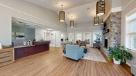 Located at 2451 Oakwood Hills Dr in Mechanicsburg, PA, this single-story community clubhouse offers a thoughtfully designed layout spanning a generous footprint across multiple functional areas. The centerpiece is a grand open lobby and lounge, distinguished by soaring vaulted ceilings, warm wood-look flooring, and stylish pendant lighting. A rich wood reception desk anchors one end of the space, while a seating area with plush sofas and chairs surrounds a stone fireplace, creating a welcoming and social atmosphere. Large windows throughout the building invite abundant natural light and offer views of the surrounding greenery. The well-equipped fitness center features a full array of cardio machines — including treadmills, ellipticals, and stationary bikes — along with a dedicated free weights station, rubber flooring, and direct exterior access. A bright dining area provides multiple seating arrangements on wood-look flooring, with easy outdoor access through glass panel doors. The kitchen is outfitted with rich wood cabinetry, granite countertops, a dishwasher, and a full-size refrigerator, ideal for community gatherings. A private meeting room with carpeted flooring and a round table offers a quiet space for consultations or small group activities. A sunlit media room with a wall-mounted TV and large windows completes the facility.