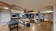 Located at 6301 Overbrook Avenue in Philadelphia, PA, this thoughtfully designed single-floor space offers a dynamic blend of lounge, work, and recreational areas. The open living area serves as the heart of the space, featuring warm wood-look tile flooring, exposed dark ceiling beams, and a striking mix of gray and teal accent walls adorned with rustic reclaimed wood paneling. A comfortable seating arrangement with modern gray sofas centers around a sleek white coffee table, complemented by a wall-mounted flat-screen TV and a bold Coca-Cola vending machine that adds a playful touch. A dedicated workstation area features iMac computers along a clean white desk, while a large communal table with Tulip-style chairs sits beneath an artistic bamboo ceiling installation and pendant lighting, creating an inspiring collaborative zone. A foosball table adds recreational flair to the space. A separate private room provides a quieter setting, equipped with a round white table, seating, and a large whiteboard — ideal for focused meetings or breakout sessions. An electric fireplace set against the teal feature wall adds warmth and character. Track lighting throughout ensures the space is well-lit, while small windows allow in natural light. The overall ambiance is modern, energetic, and versatile.