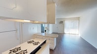 This single-floor residence offers a practical and functional layout centered around an open-concept design that seamlessly connects the kitchen and living areas. The kitchen is outfitted with white cabinetry that extends to the ceiling, maximizing storage capacity, and features a gas cooktop set into a light-toned countertop. A peninsula-style counter provides additional workspace and serves as a natural dividing point between the kitchen and the adjoining living space. The textured ceiling adds a classic residential character throughout the interior. The flooring transitions to warm wood-tone laminate or hardwood in the living area, lending a cohesive and welcoming feel to the space. The living room benefits from natural light streaming through a sliding glass door dressed with vertical blinds, which also provides direct access to the outdoors. A wall-mounted HVAC unit ensures climate control convenience. The overall palette of whites and neutral tones creates a clean, airy ambiance, making the space feel open and easy to personalize. The straightforward single-floor layout offers ease of navigation and accessibility throughout.