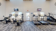 This modern fitness center offers a comprehensive workout environment housed within a single, open-plan space. The room immediately impresses with its soaring ceilings, which are accented by a decorative wood beam detail and recessed lighting, lending the space a bright and airy feel. Large windows along the perimeter allow ample natural light to flood the interior and provide views of the surrounding outdoor area, including what appears to be a pool and courtyard.

The flooring throughout is a patterned modular carpet tile in neutral tones of grey and blue, providing both comfort and durability. The walls are finished in a clean white, enhancing the sense of openness.

The fitness center is thoroughly equipped to support a variety of workout routines. Cardio equipment includes multiple treadmills, elliptical trainers, stationary and cycle bikes, and a rowing machine. For strength and functional training, the space features a cable/multi-use machine, a weight bench, free weights, and additional accessories on dedicated shelving. A floor-to-ceiling mirror panel assists with form and adds depth to the room.

Multiple wall-mounted flat-screen televisions are positioned throughout the space for entertainment during workouts. Industrial-style wall fans provide additional ventilation. A glass door opens directly to the outdoor amenity area, further connecting the fitness center to the broader community.