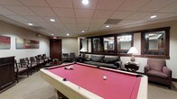 This single-floor recreational lounge is thoughtfully designed to offer a welcoming and functional space for leisure and social gatherings. At the heart of the room stands a full-size billiards table dressed in striking red felt, serving as the focal point of the space. The room is furnished with plush, dark upholstered sofas and accent chairs, providing comfortable seating areas for guests to relax and unwind. Warm table lamps contribute a soft, inviting ambiance that complements the recessed ceiling lighting set within a classic drop-tile ceiling. A wine or beverage cooler is tucked neatly into the seating area, adding a convenient hospitality touch. A set of large interior windows with dark wood framing opens a sightline into an adjacent room, where a chandelier is visible, lending an air of elegance. Dark wood doors and trim throughout the space provide a refined, cohesive aesthetic. The neutral carpeted flooring and warm-toned walls further enhance the cozy, upscale atmosphere, making this lounge a versatile and inviting retreat for recreation and socializing alike.