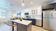 This thoughtfully designed 1-bedroom, 1-bathroom unit offers a modern open-concept layout that efficiently maximizes every square foot. The kitchen is a standout feature, boasting two-tone cabinetry with crisp white upper cabinets and deep navy lower cabinets, a white subway tile backsplash, and a full suite of stainless steel appliances including a range, microwave, dishwasher, and refrigerator. A central island with bar seating and a pendant light overhead makes the kitchen both functional and social, flowing seamlessly into the living area. The living room features light wood-look flooring, a ceiling fan, and large windows dressed with horizontal blinds that invite natural light. The bedroom is a calm retreat, featuring a window with natural light, light wood flooring, and a ceiling fan. A practical hallway area connects the bedroom to a dedicated closet space and an in-unit stacked washer and dryer — a notable convenience. The bathroom is clean and straightforward with a dark vanity complementing the unit's modern palette. Throughout the space, white walls and consistent light wood flooring create a cohesive, airy ambiance well-suited for comfortable everyday living.