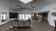 Located at 201 Genung St in Middletown, New York, this single-floor space offers an open, welcoming layout designed for comfortable gathering and daily use. The main lounge area is anchored by soaring vaulted ceilings fitted with dual ceiling fans, creating excellent airflow and a sense of generous volume throughout. A large skylight adds to the brightness of the space, complementing the abundant natural light that pours in through a row of French doors along the exterior wall. These glass-paned doors not only illuminate the interior but also provide easy access to the outdoors. The lounge is furnished with a curved sofa and patterned area rug, lending the space a warm and inviting atmosphere. A large flat-screen television is mounted on an accent wall, adding a modern entertainment element. Adjacent to the lounge, a full kitchen is visible with a breakfast bar, pendant lighting, warm wood cabinetry, and stainless steel appliances, making it well-equipped for hosting and everyday needs. Bar-height seating at the counter encourages casual dining and socializing. The neutral color palette, carpeted flooring in the lounge, and the seamless flow between living and kitchen areas contribute to a cohesive, functional, and comfortable environment throughout.