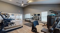 Located at 35 Lucy Ridge Rd in Delaware, Ohio, this fitness center offers a thoughtfully designed workout space that maximizes functionality and comfort. The room features striking vaulted ceilings that soar to impressive heights, creating an open, airy atmosphere enhanced by large transom windows that flood the space with natural light. A ceiling fan provides additional air circulation throughout.

The flooring consists of durable hardwood-style planks, complemented by protective rubber exercise mats in designated workout zones. Full-length wall mirrors are strategically positioned to expand the visual space and assist users in monitoring their form during exercise.

The facility is comprehensively equipped with a wide variety of fitness equipment, including multiple treadmills, an elliptical trainer, a stationary bike, a multi-station cable and strength machine, an adjustable weight bench, and a full dumbbell rack with free weights ranging across multiple sizes. Resistance bands and a step platform are also available, catering to diverse workout preferences.

Multiple wall-mounted flat-screen televisions provide entertainment during workouts. The space is accessed through elegant frosted-glass double doors, adding a polished, modern aesthetic. A designated area for dirty towels reflects thoughtful attention to facility cleanliness and user convenience.