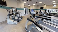 The fitness center at District At Hurstbourne offers a spacious, thoughtfully designed workout facility that caters to a wide range of exercise needs. The interior is defined by a clean, neutral color palette with a distinctive teal accent band running along the walls, giving the space a fresh and energetic atmosphere. Light-toned carpet tile flooring spans the entire area, providing both comfort and durability underfoot.

Large windows line multiple walls of the facility, allowing abundant natural light to fill the space while offering views of the surrounding landscaped grounds. Multiple ceiling fans are distributed throughout, complementing the recessed panel lighting to maintain a comfortable environment during workouts.

The cardio section is well-stocked with a row of treadmills, elliptical trainers, and stair climbers from True and Precor, offering residents a variety of aerobic options. A dedicated strength training area features a multi-station cable machine and weight bench, while a dumbbell rack with a full range of free weights is positioned against a large window. Medicine balls mounted on a wall rack provide additional functional training options.

Wall-mounted televisions are positioned for visibility throughout the space. A glass-paneled entry door with views to the exterior makes for an inviting entrance. The overall layout is open and uncluttered, ensuring ease of movement and a motivating environment for daily fitness routines.