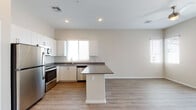 This single-floor unit features a clean, contemporary design with a neutral color palette and consistent light wood-look flooring that flows throughout the space. The open-concept kitchen and living area form the heart of the unit, where white flat-panel cabinetry extends to the ceiling, providing ample storage. Dark quartz-style countertops contrast crisply against the white cabinets, and a peninsula extends outward to define the kitchen space while offering additional prep and seating area. A full suite of stainless steel appliances—including a refrigerator, range, microwave, and dishwasher—adds a modern, functional finish. A window above the sink brings in natural light.

The adjoining living area is generously proportioned with high, angled ceilings and recessed lighting, creating an airy atmosphere. Bedrooms maintain the same light flooring and neutral warm-toned walls; one bedroom features a sliding door with vertical blinds that opens to an outdoor area, along with a corner window for additional natural light. The bathroom showcases a wide vanity with dark countertops, white lower cabinetry with brushed hardware, and a large mirror. The overall ambiance is bright, modern, and move-in ready.