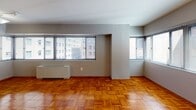 Located on the fifth floor at 522 21st St NW in Washington, DC, this apartment offers a well-organized single-floor layout with a clean, understated aesthetic throughout. The living area is a standout feature, characterized by warm-toned parquet wood flooring laid in a geometric pattern that adds timeless character to the space. Multiple large windows fitted with horizontal blinds line the walls, allowing generous natural light to filter in while framing views of the surrounding urban cityscape. A wall-mounted HVAC unit provides climate control directly within the main living space. The bedroom is a quiet, well-proportioned room that continues the parquet flooring and benefits from its own bank of windows, offering additional natural light. A structural column detail and open doorway connect the bedroom to the rest of the unit, with a closet providing practical storage. The bathroom showcases a classic mid-century design with crisp white subway tile walls accented by bold black tile borders, a combined tub and shower with a curtain rod, a pedestal-style sink, and a large mirror. The neutral gray wall tones throughout the unit create a versatile backdrop, while the consistent parquet flooring unifies the space. Situated in the heart of Washington, DC, this apartment is ideally positioned for access to the city's amenities.