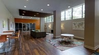 This single-floor space features an open-plan layout with rich hardwood flooring throughout, creating a warm and welcoming atmosphere. The design incorporates high ceilings with recessed lighting and large clerestory windows along the upper walls, flooding the interior with natural light and offering views of surrounding greenery. A bold orange accent wall near the entrance adds a striking pop of colour, complementing the contemporary aesthetic. The lounge area is anchored by a dark upholstered sectional sofa arranged atop a colourful patterned area rug, providing a comfortable setting for relaxed gatherings. Adjacent to this, a round white table with modern chair seating offers a practical space for casual meetings or individual use. Floor lamps positioned throughout the room contribute to a warm, layered lighting scheme. The entryway features glass-panelled doors that enhance the sense of openness and connectivity to the exterior. Artwork and decorative elements on the walls add personality and visual interest. The overall ambiance is modern and functional, balancing vibrant design choices with a clean, inviting atmosphere suited to community interaction.