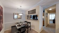 This single-story residence showcases a thoughtfully designed open-concept layout that seamlessly connects the dining area to the kitchen. The dining space is anchored by a stylish pendant light fixture suspended from a textured ceiling, casting warm illumination over the glass-top dining table and dark wood chairs below. A decorative wall mirror enhances the sense of openness, while a floating shelf on the dividing wall provides a functional display area. The partial wall between the dining room and kitchen features a pass-through opening, allowing natural visibility between the two spaces and contributing to an airy, connected feel. The kitchen, visible through the pass-through, is equipped with warm wood cabinetry and appears well-appointed for everyday use. Neutral beige walls and light-toned carpeting throughout the dining area create a cohesive, understated palette that complements the darker furniture accents. Crown molding details and clean white baseboards add a refined finishing touch to the interior. Overall, the space strikes a balance between practicality and comfort, making it well-suited for both everyday living and casual entertaining.
