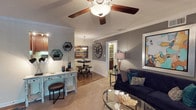 This single-floor space offers a welcoming and elegantly appointed interior designed for both relaxation and social interaction. The open-concept layout allows for a seamless flow between seating and dining areas, creating a versatile environment suited to a variety of uses.

The main living area is anchored by a rich, dark wood console sideboard with decorative detailing, above which a wall-mounted flat-screen television serves as a focal point. Warm earth-toned walls in deep brown and gold hues create an inviting ambiance, complemented by classic table lamps that cast a soft, ambient glow throughout the space.

Comfortable upholstered seating in golden tones is arranged around a glass-topped coffee table atop a decorative area rug, providing an ideal space for conversation or leisure. Hardwood flooring in a warm honey tone extends throughout, adding to the overall sense of warmth and cohesion.

Recessed lighting in the white ceiling ensures consistent, well-distributed illumination across the entire floor. Toward the rear, natural light filters in through windows, brightening a secondary dining area furnished with additional seating. Lush potted plants are thoughtfully placed throughout, introducing a refreshing natural element. The overall ambiance is classic, comfortable, and carefully curated for communal living.