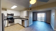 This single-story unit offers a well-organized layout with distinct living, sleeping, and functional zones. The kitchen is a standout feature, equipped with a full suite of stainless steel appliances including a gas range, microwave, refrigerator, and dishwasher. Crisp white cabinetry pairs with dark granite-style countertops, while light wood-look flooring and a recessed ceiling light panel keep the space bright and practical. A pendant light fixture with a gold-toned accent adds a subtle decorative touch overhead.

The kitchen opens seamlessly into the living area, which continues the wood-look flooring and features a ceiling fan. A sliding glass door at the far end of the living space allows natural light to filter in and provides access to an outdoor area. The transition from the kitchen to the living space is marked by a subtle change in flooring tone, adding visual definition between zones.

The bedroom offers plush carpeting, neutral walls, a textured ceiling, and two windows that bring in ample natural light. The bathrooms are finished with white cabinetry, granite-style countertops, and tub/shower combinations, maintaining a clean and consistent aesthetic throughout the unit.