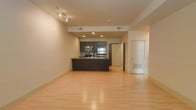 This single-floor space showcases a thoughtfully designed open-concept layout that seamlessly blends the living and kitchen areas into one cohesive environment. The kitchen is a standout feature, outfitted with dark espresso cabinetry, stainless steel appliances including a refrigerator, microwave, and range, as well as a peninsula countertop that doubles as a functional workspace or casual dining surface. Recessed lighting and track lighting fixtures illuminate the space efficiently, complementing the neutral, light-toned walls that lend a clean and airy feel. Light hardwood flooring extends throughout, adding warmth and continuity to the open floor plan. The ceiling features integrated HVAC vents, ensuring a streamlined and uncluttered overhead aesthetic. The generous open area of the living space offers flexibility for a variety of furniture arrangements. Doorways leading to additional rooms or utility areas are visible, suggesting a practical and well-organized floor plan. Overall, the space conveys a modern, minimalist ambiance that is both functional and inviting, ideal for those who appreciate contemporary design and efficient use of space.