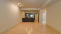 This single-floor space showcases a thoughtfully designed open-concept layout that seamlessly blends the living and kitchen areas into one cohesive environment. The kitchen is a standout feature, outfitted with dark espresso cabinetry, stainless steel appliances including a refrigerator, microwave, and range, as well as a peninsula countertop that doubles as a functional workspace or casual dining surface. Recessed lighting and track lighting fixtures illuminate the space efficiently, complementing the neutral, light-toned walls that lend a clean and airy feel. Light hardwood flooring extends throughout, adding warmth and continuity to the open floor plan. The ceiling features integrated HVAC vents, ensuring a streamlined and uncluttered overhead aesthetic. The generous open area of the living space offers flexibility for a variety of furniture arrangements. Doorways leading to additional rooms or utility areas are visible, suggesting a practical and well-organized floor plan. Overall, the space conveys a modern, minimalist ambiance that is both functional and inviting, ideal for those who appreciate contemporary design and efficient use of space.
