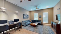 Located at 833 E Magnolia Dr in Tallahassee, Florida, this thoughtfully designed single-floor space blends comfort and functionality in a contemporary setting. The main lounge area is anchored by a generously sized blue velvet sectional sofa paired with a warm wood coffee table, all set atop a patterned area rug that adds texture and depth to the room. Neutral taupe walls provide a versatile backdrop, while large windows with white blinds invite ample natural light throughout. Ceiling fans with recessed lighting maintain a comfortable and well-lit atmosphere. A herringbone-patterned wood credenza with a wall-mounted flat-screen television serves as a refined focal point, complemented by large-scale abstract artwork adorning the walls that adds a curated, gallery-like character to the space. Light hardwood flooring runs continuously throughout, lending warmth and cohesion. A dedicated workstation area features a dark wood desk with dual monitors, a printer, and modern pendant lighting overhead — ideal for productivity needs. French glass doors connect the work area to the broader space, maintaining an open and airy flow. The overall ambiance is modern and polished, making this space well-suited for both relaxation and focused work.