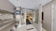 This single-floor hallway serves as a fully functional residential lobby and corridor, offering a generous and well-organized layout. The space is defined by its classic terrazzo flooring, featuring a speckled black-and-white pattern with dark inlay strips that lend a timeless, polished character throughout.

The entry vestibule is secured with glass-paneled doors and a key fob or intercom access system, providing a clear delineation between the exterior and interior. A built-in mailbox bank is conveniently located near the entrance, accompanied by a dedicated parcel shelf for resident use.

Moving deeper into the lobby, the space opens into a welcoming seating area furnished with leather armchairs and a central side table, creating a comfortable communal gathering point. A large mirror panel enhances the sense of openness in this area. The elevator is prominently positioned nearby, framed by a distinctive dark mosaic tile accent wall that adds a bold visual element.

The corridor extends toward individual unit doors, including numbered suites, with clean white walls and recessed and flush-mount ceiling lighting providing consistent, even illumination throughout. The textured ceiling and understated neutral palette maintain a functional yet refined ambiance across the entire space.